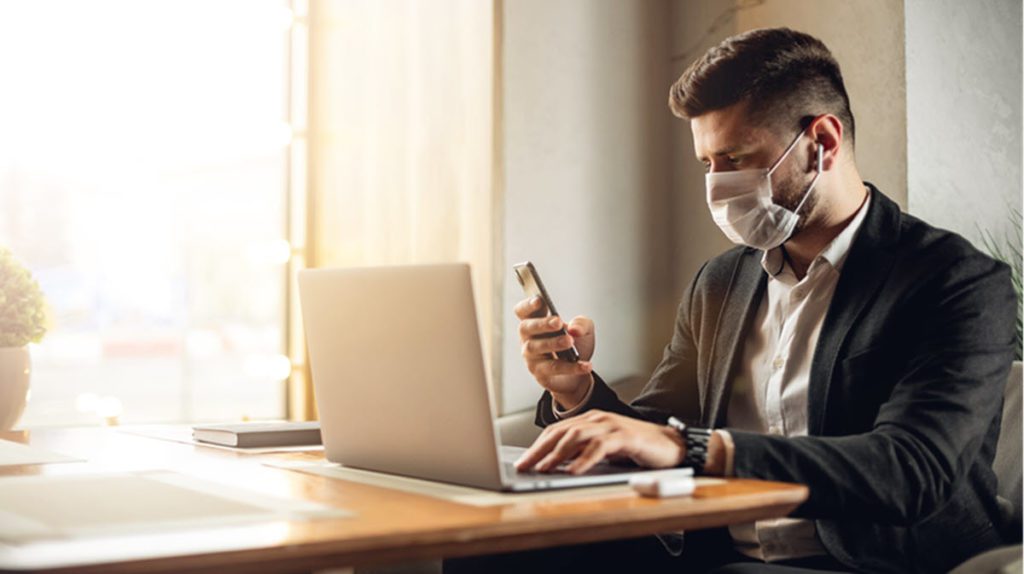 Person mit Mundschutzmaske vor dem Laptop, Smartphone in der Hand